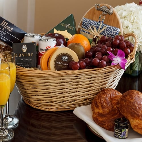 A wicker basket overflows with gourmet foods, including cheeses, fruits, and caviar. Nearby, champagne and orange juice rest beside croissants on a table in Key West. Text includes: