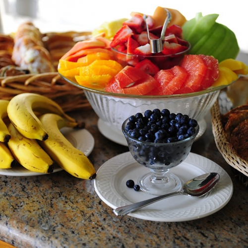 Bowl of assorted fruits, next to a tray full of different breads and pastries, a plate of bananas, and bowl of blueberries.