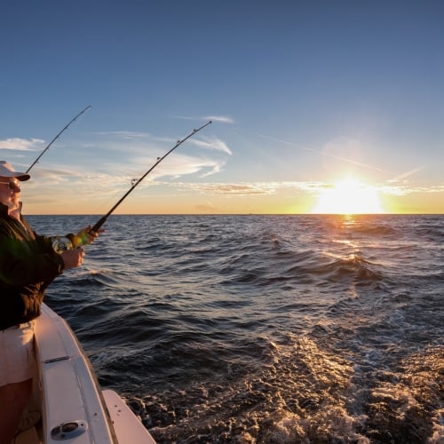 A man fishes from a boat, holding two rods as the sun sets, illuminating the ocean waves near Key West.