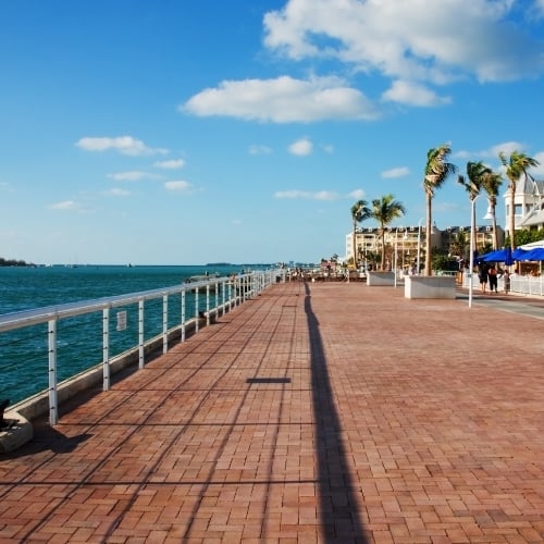 A long brick walkway stretches by calm turquoise waters with railings, palm trees, and nearby benches. People stroll near waterfront cafes with blue umbrellas in Key West, under a bright blue sky.