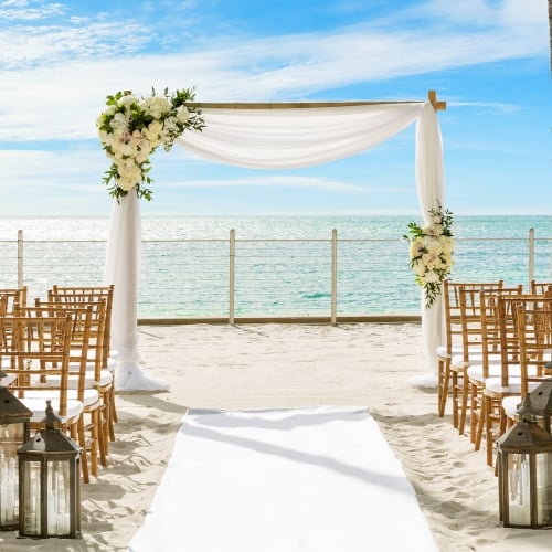 A floral arch stands on a sandy beach, surrounded by chairs and lanterns in Key West. The ocean and palm trees create a serene wedding setting under a bright blue sky.