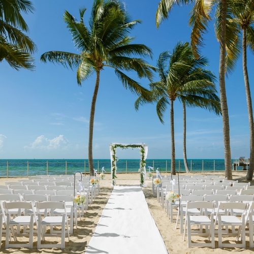 Wedding setup on the beach, including rows of white chairs, a decorated archway, and palm trees.