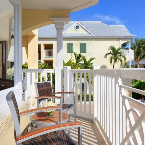 Two chairs and a small table on a balcony, facing lush palm trees and pastel-colored buildings under a clear blue sky in Key West.