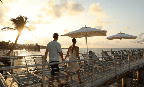 Couple walking along the pier at sunset lined with lounge chairs and beach umbrellas.