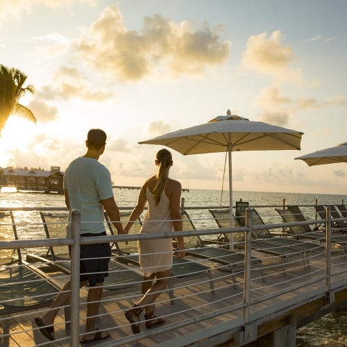 Couple walking along the pier at sunset with lounge chairs arranged in a row and white beach umbrellas.