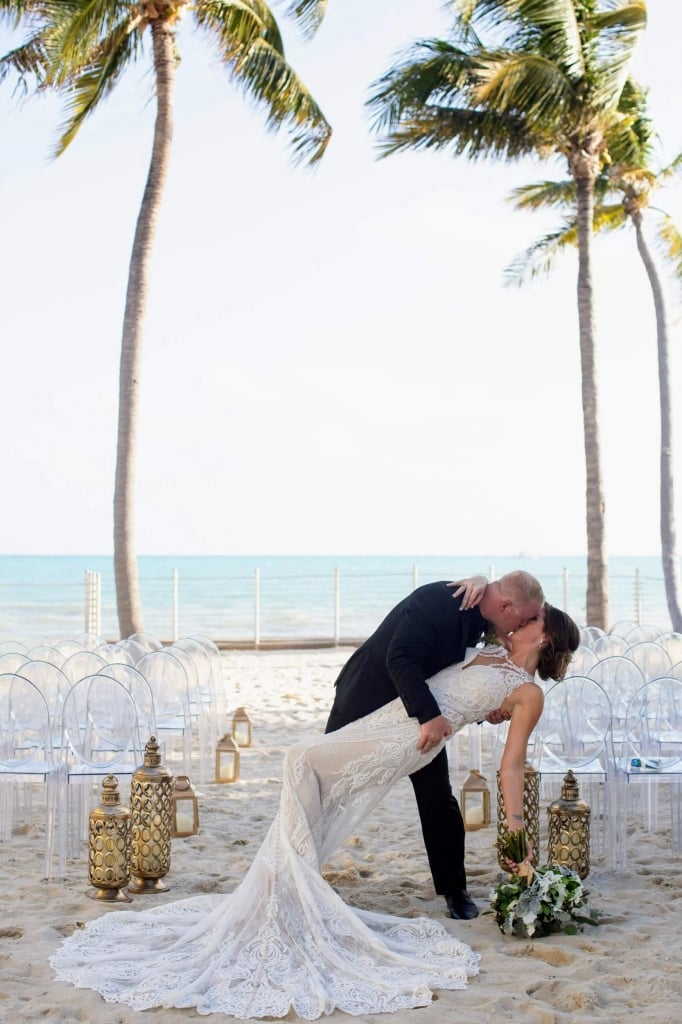 A husband dips his new bride while kissing her on the beach with clear chairs and palm trees in the background on their wedding day.