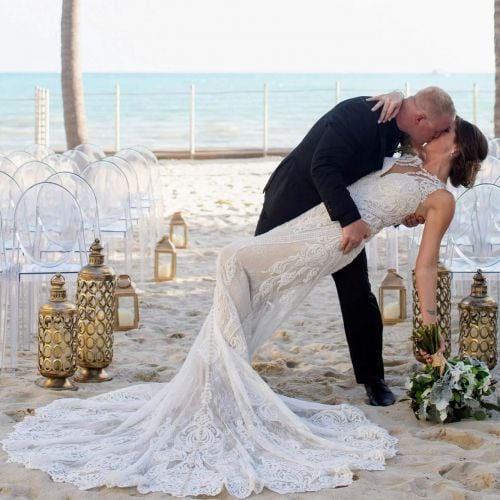 A husband dips his new bride while kissing her on the beach with clear chairs and palm trees in the background on their wedding day.