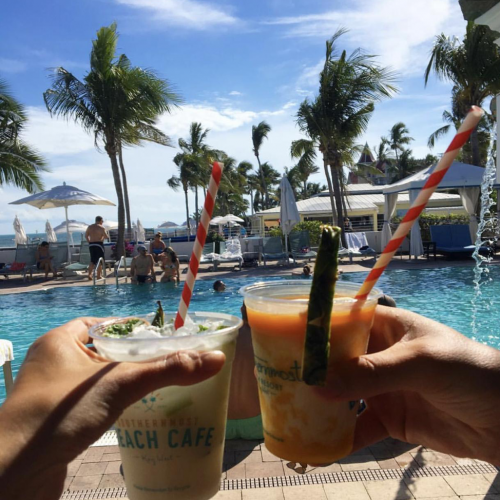 Two drinks with Beach Cage logo are held side-by-side on the beach with garnishes and red and white straws.