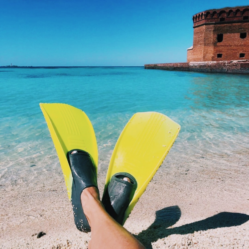 A view of man's feet in flippers as he sits in the sand looking out to the ocean.