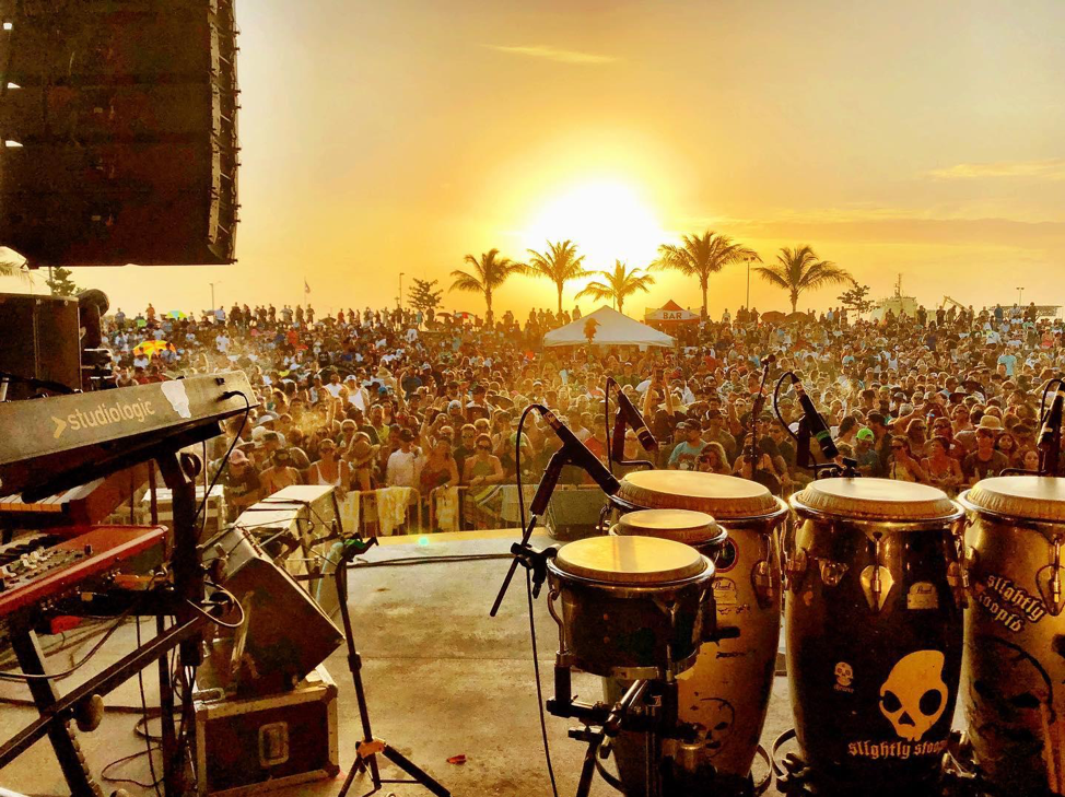 View of instruments on an outdoor stage looking into a large crowd at sunset.