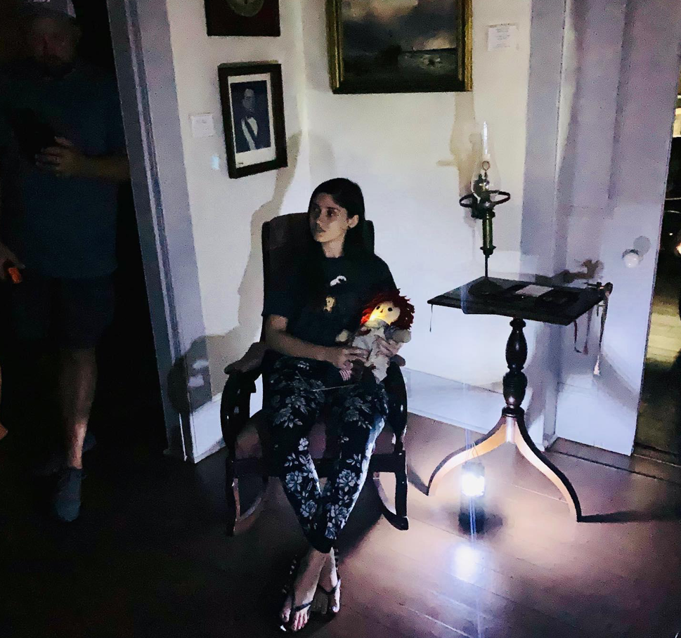 Young woman sits in a chair while holding a doll in a dimly lit room as part of the Haunted Key West Tour.