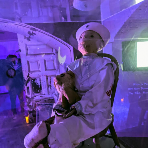 A vintage doll in a sailor outfit sits on a chair holding a stuffed toy. The display is in a glass case with purple lighting, located in Key West.