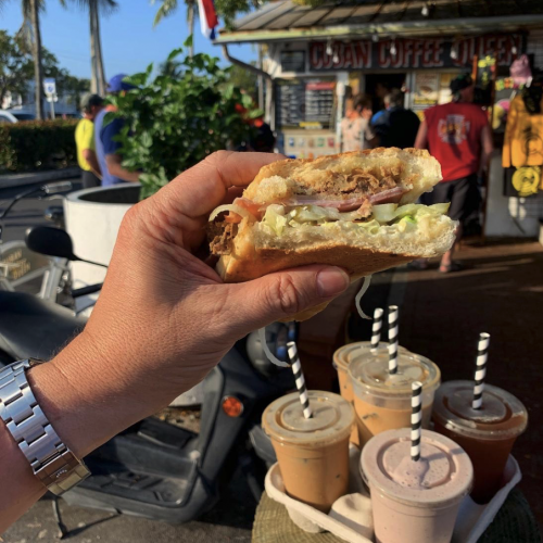 A hand holds a bitten sandwich filled with lettuce and meat in front of a food stand labeled