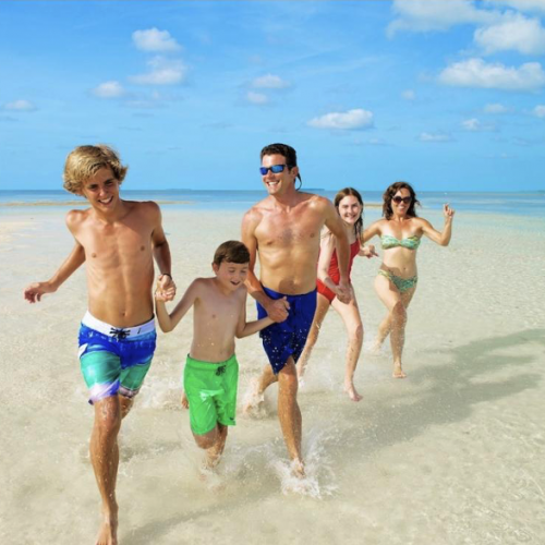 A group of five people is joyfully running through shallow water on a sunny beach in Key West, with clear blue skies and an expansive seascape as the backdrop.