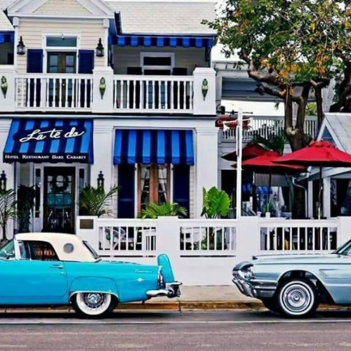 Two vintage cars are parked in front of La Te Da Hotel, Restaurant, Bar, and Cabaret, featuring blue and white awnings and red umbrellas in Key West, providing a charming, classic atmosphere.