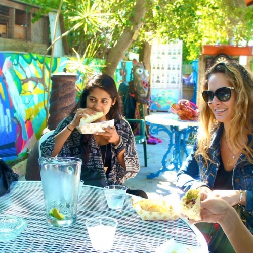 Women are eating lunch and enjoying conversation at an outdoor table in The Keys. The setting features vibrant murals and lush greenery, creating a lively and colorful atmosphere.