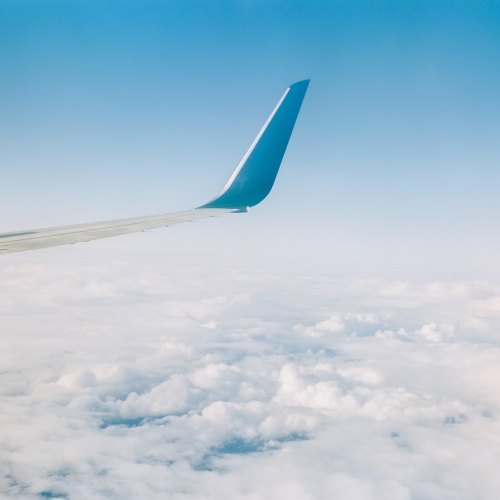 Airplane wing slicing through fluffy white clouds under a clear blue sky, viewed from a window. Geo-location: Key West or The Keys.