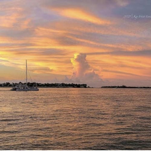 A sailboat drifts across calm waters during sunset near Key West, with a silhouetted island in the distance under a colorful, cloud-filled sky.