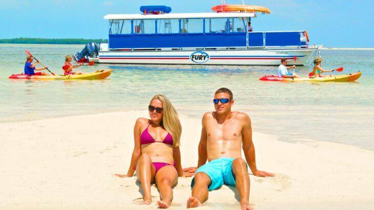 Man and woman sitting on a beach with a boat and kayakers in the background