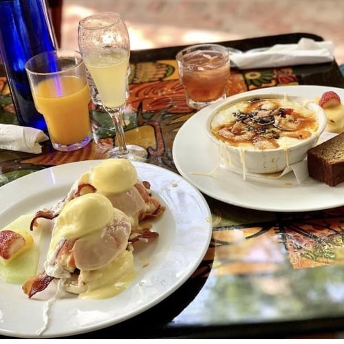 A table in Key West features eggs Benedict on an artful plate, next to a dish with baked cheese, toast, and fruit. Surrounding drinks include orange juice, mimosa, and a cocktail.