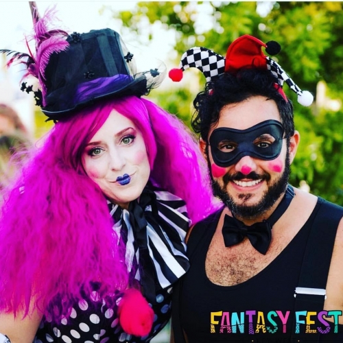 Two people in vibrant costumes pose for a photo. One wears a purple wig and top hat, the other a jester hat with a black mask.