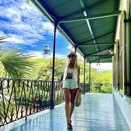 A person walks along a turquoise-painted veranda. They're wearing summer clothes, holding a straw bag. A lighthouse and lush greenery appear under a bright blue sky in Key West.