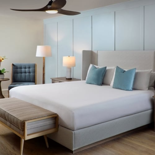 A neatly made bed with blue pillows sits in a modern, well-lit room featuring chairs, lamps, and a fan, suggesting a relaxing atmosphere in Key West.