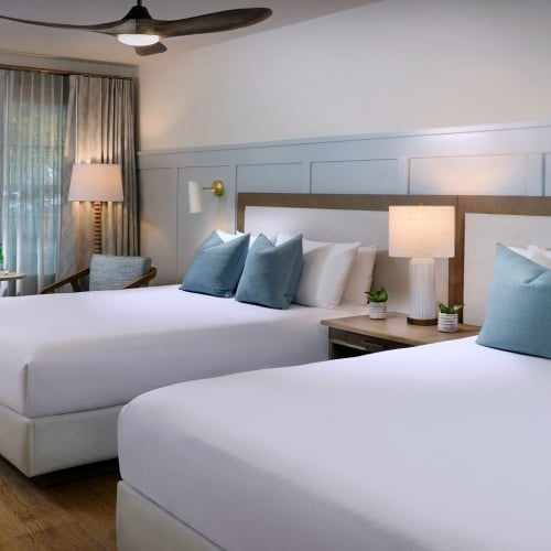 Two neatly made beds feature blue accent pillows, situated in a bright, airy room with wooden floors and a ceiling fan. A window with sheer curtains and a lamp create a cozy ambiance in Key West.