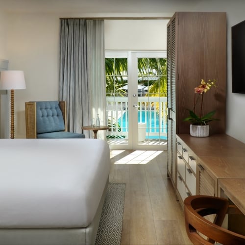A cozy hotel room in Key West features a bed with blue pillows, a wooden desk, and a large TV. French doors lead to a balcony overlooking a palm tree and pool.