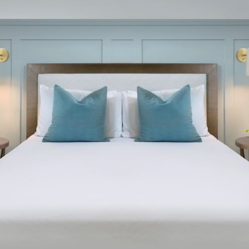A neatly made bed with two blue pillows rests against a light blue paneled wall, flanked by two wooden nightstands with wall-mounted lamps and a small plant, in a Key West setting.