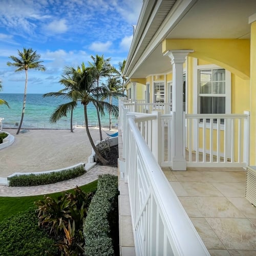 Yellow beachside buildings overlook a sandy path leading to the ocean, edged by palm trees, in a tropical Key West setting under a partly cloudy blue sky.