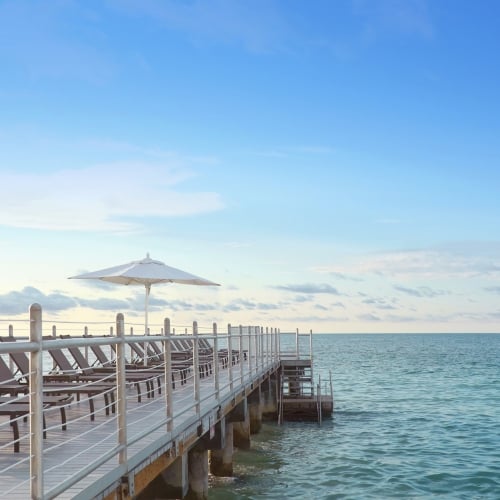 Lounge chairs and a white umbrella rest on a wooden pier extending over calm ocean waters, under a clear blue sky in Key West.