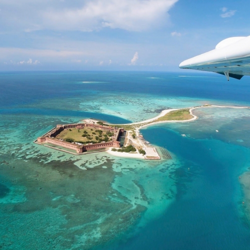 Aerial view of Fort Jefferson, a large brick structure on a small island. Turquoise waters surround the fort, with a few boats nearby, underneath a plane wing. Located in The Keys.