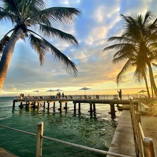 Palm trees frame a wooden pier where people are gathered, illuminated by a warm sunset over the ocean. Located in Key West, the scene exudes a tropical, serene ambiance.