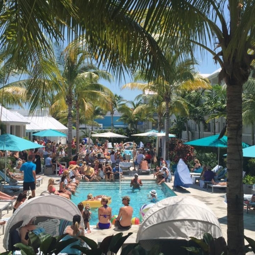 A crowded pool area features people lounging under umbrellas and swimming. Palm trees and tropical foliage provide shade, creating a lively resort atmosphere in Key West.