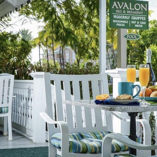 A table set with breakfast items such as orange juice, a muffin, and fruit sits on a porch surrounded by white chairs in Key West. A sign reads,