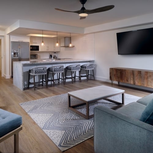 A modern living room in Key West, featuring a blue sofa facing a wall-mounted TV. A wooden coffee table sits on a patterned rug. Behind, a sleek kitchen with bar stools and pendant lights creates a stylish, open-concept space.
