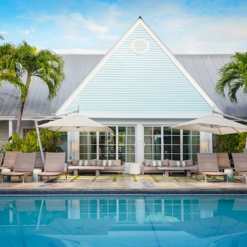 A pool reflects lounge chairs under umbrellas, positioned in front of a pastel blue building surrounded by palm trees. Clear skies enhance the tranquil setting in Key West.