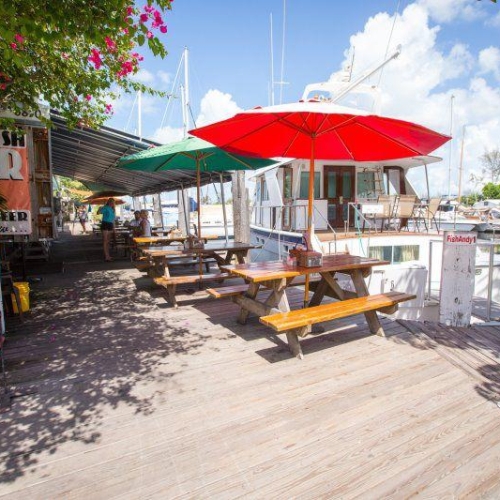 Picnic tables sit under colorful umbrellas on a wooden deck beside a marina, with boats docked in the background. Sunny weather and tropical plants enhance the Key West coastal atmosphere.