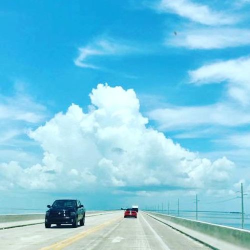 Two cars drive on a concrete bridge, stretching over water, under a bright blue sky with fluffy clouds in Key West.