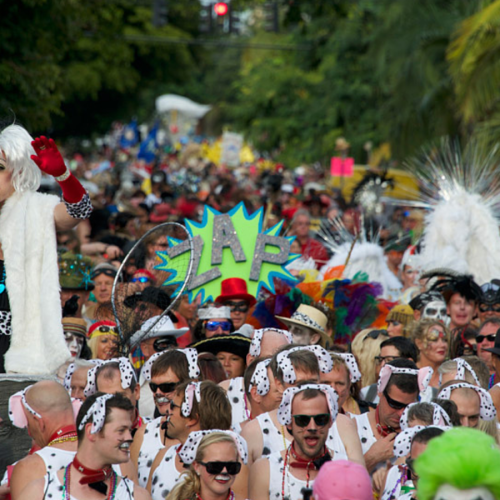 Performer stands on a parade float, speaking into a microphone, surrounded by a festive crowd in costumes, including Dalmatian outfits. A