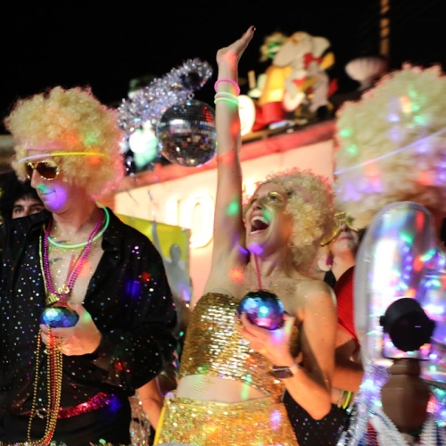 People in colorful, disco-themed costumes dance enthusiastically, holding shiny objects under vibrant party lights. A neon sign reads