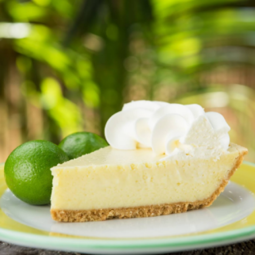 A slice of Key lime pie with whipped cream sits on a yellow-green plate beside whole limes, set against a blurred background of lush green foliage in The Keys.