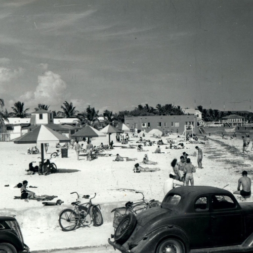People are relaxing and socializing on a sandy beach with umbrellas and bicycles nearby, bordered by palm trees and old cars under a clear sky in Key West.