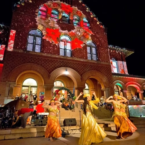 Dancers in yellow and orange costumes perform energetically on steps. Spectators watch in front of a decorated building featuring festive lights and projections. Nighttime festival setting in Key West.