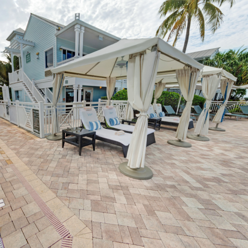 Poolside cabanas with lounge chairs under white canopies provide relaxation beside a clean, inviting pool. Tropical palm trees and blue buildings enhance the leisurely atmosphere in Key West.