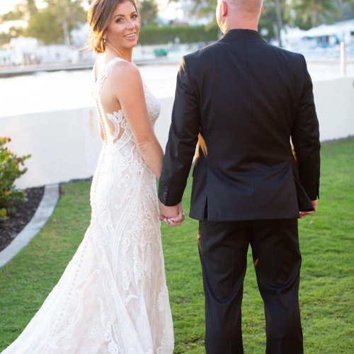 A couple holds hands; the woman in a lace wedding dress looks back, smiling. They're standing on grass near the waterfront, with palm trees and sunlight in Key West.