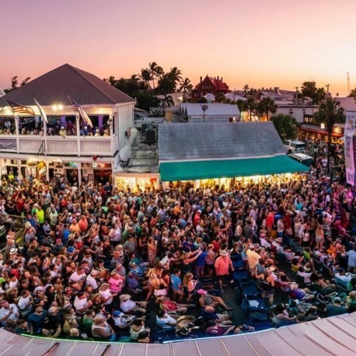 A large crowd watches a band performing on an outdoor stage during sunset in Key West. People fill the street, surrounded by illuminated shops and buildings, creating a lively atmosphere.