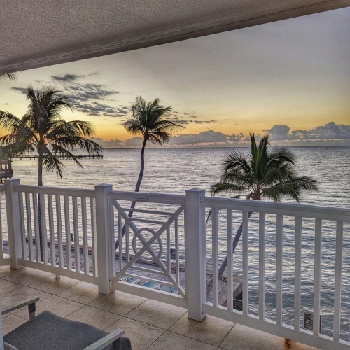 A white balcony overlooks the ocean at sunset, with palm trees gently swaying in the breeze. The Keys provide a tropical backdrop, enhancing the serene coastal view.