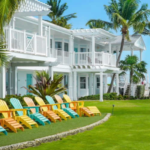 Colorful Adirondack chairs are aligned on a lush lawn facing a white, two-story beach house. Palm trees sway nearby, with loungers and a cornhole game set up. Location: Key West.